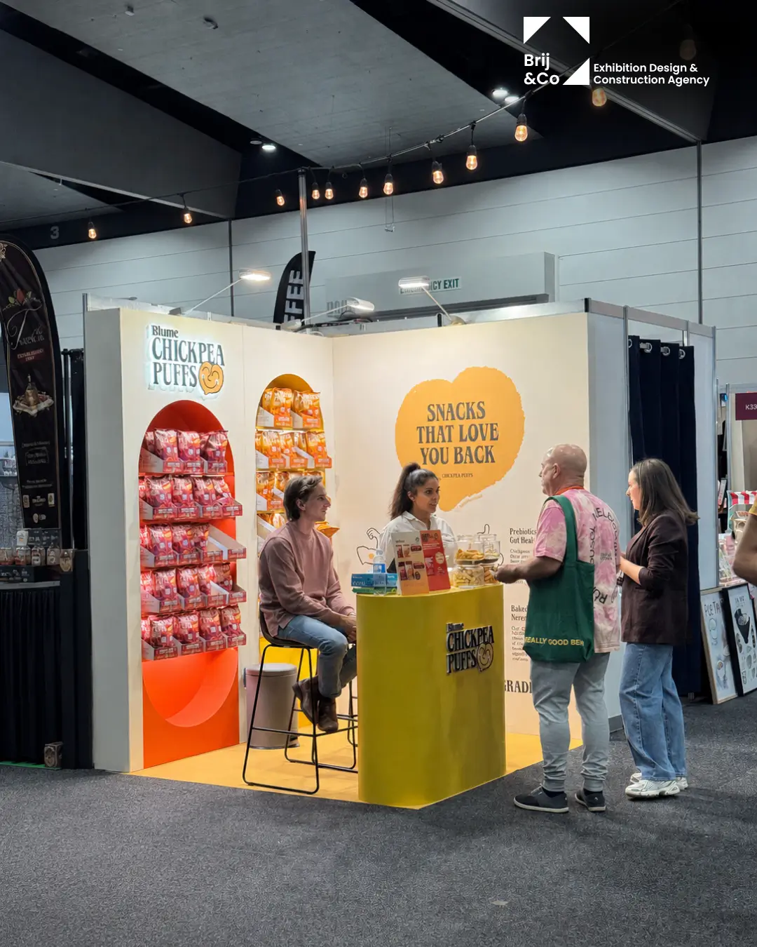 Blume Chickpea Puffs exhibition stand with visitors at the yellow counter